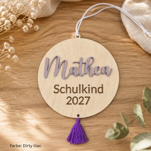 May include: A circular light-colored wooden ornament featuring the name Mathea in raised lilac script lettering. Below the name, the text Schulkind 2027 is engraved in a dark font. A white string is attached to the top, and a lilac tassel hangs below.