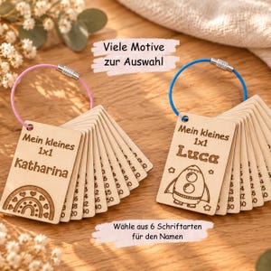 Könnte beinhalten: Zwei Sets rechteckiger Multiplikationskarten aus Holz an Metallringen. Ein Set hat einen rosa Ring und ein Regenbogenmotiv mit dem Namen Katharina, das andere einen blauen Ring und ein Raketenmotiv mit dem Namen Luca. Beide tragen den Text Mein kleines 1x1.