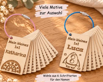 Può includere: Due set di schede di moltiplicazione rettangolari in legno su anelli di metallo. Un set ha un anello rosa e un design arcobaleno con il nome Katharina, l'altro un anello blu e un design a razzo con il nome Luca. Entrambi riportano il testo Mein kleines 1x1.