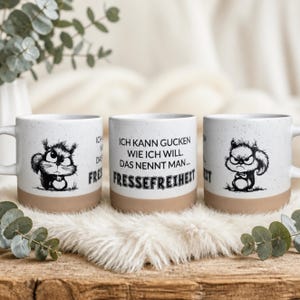 Könnte beinhalten: Ein weißer Keramikbecher mit Sprenkel-Optik und beigem Boden. Der Becher zeigt eine schwarze Tuschezeichnung eines mürrischen Eichhörnchens und den Text: ICH KANN GUCKEN WIE ICH WILL. DAS NENNT MAN FRESSEFREIHEIT. Das Motiv ist beidseitig abgebildet.