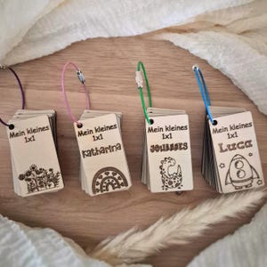 May include: Four sets of rectangular wooden tags held together by colored metal rings. Each tag features the laser-engraved text "Mein kleines 1x1" and a unique illustration: flowers, a rainbow, a dinosaur, or a rocket, with names Katharina, Johannes, and Luca.