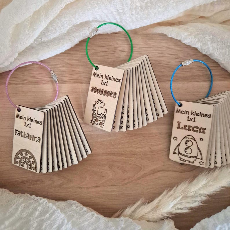 May include: Three sets of wooden multiplication flashcards on metal rings. Each set features a name, the text Mein kleines 1x1, and a graphic: a rainbow for Katharina, a dinosaur for Johannes, and a rocket ship for Luca. The cards are light wood.