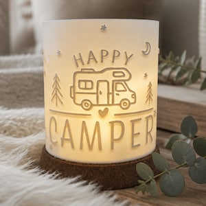 May include: A cylindrical white illuminated lamp with a cork base featuring a camper van graphic, a small heart, pine trees, stars, and a crescent moon. The text HAPPY CAMPER is embossed on the front. The lamp emits a warm glow from within.