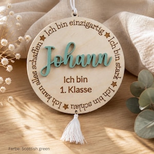 May include: A circular wooden ornament featuring the name Johann in raised teal script. The text Ich bin einzigartig, Ich bin stark, Ich bin schlau, Ich kann alles schaffen, and Ich bin 1. Klasse is engraved around the center. A white tassel hangs from the bottom.
