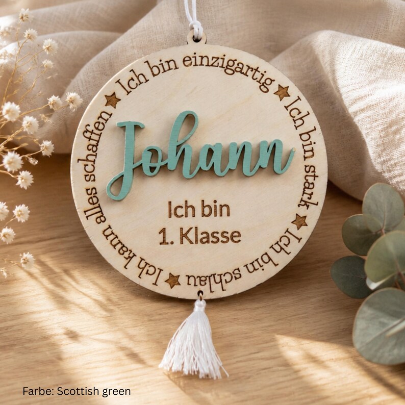 May include: A circular wooden ornament featuring the name Johann in raised teal script. The text Ich bin einzigartig, Ich bin stark, Ich bin schlau, Ich kann alles schaffen, and Ich bin 1. Klasse is engraved around the center. A white tassel hangs from the bottom.