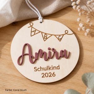 May include: A circular wooden ornament featuring a laser-engraved bunting banner with the letters A, B, and C. The name Amira is displayed in raised, coral-blush colored script lettering, followed by the engraved text Schulkind and 2026.