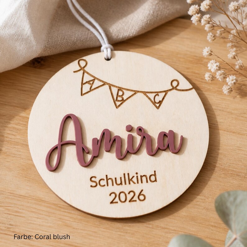 May include: A circular wooden ornament featuring a laser-engraved bunting banner with the letters A, B, and C. The name Amira is displayed in raised, coral-blush colored script lettering, followed by the engraved text Schulkind and 2026.