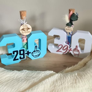 Könnte beinhalten: Zwei Geburtstagsdekorationen mit der Zahl "30" in Blau und Weiß, mit "29+" und einem Mittelfinger-Symbol. Jede hat ein Glasröhrchen mit Geld und einem "Happy Birthday"-Anhänger. Eines hat einen Holzanhänger, das andere Blumen.
