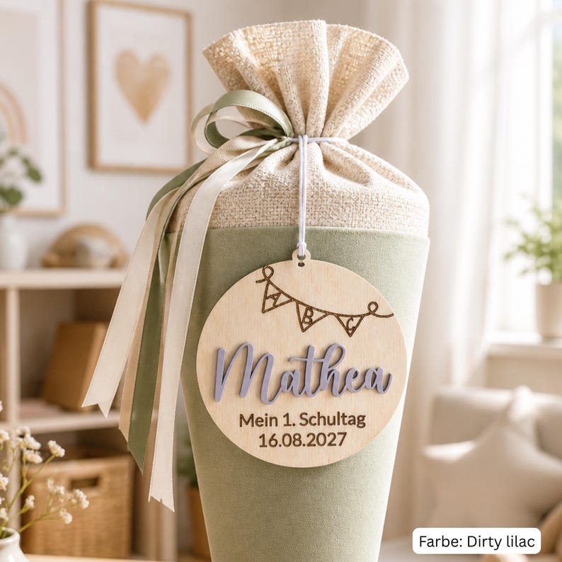 May include: A cone-shaped school gift bag in dirty lilac fabric with a beige burlap top tied with sage green and cream ribbons. A round wooden tag hangs from the front, featuring an engraved ABC bunting, the name Mathea, and the text Mein 1. Schultag 16.08.2027.