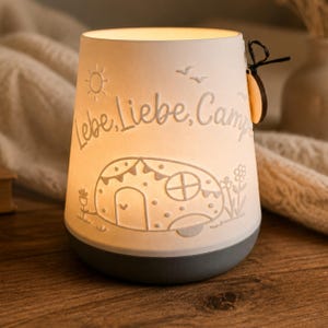 May include: A white ceramic lantern with a grey base, illuminated from within. The surface features embossed designs of a camper, flowers, a sun, and birds, with the text Lebe, Liebe, Campe. A small wooden tag is attached to the side with a black string.