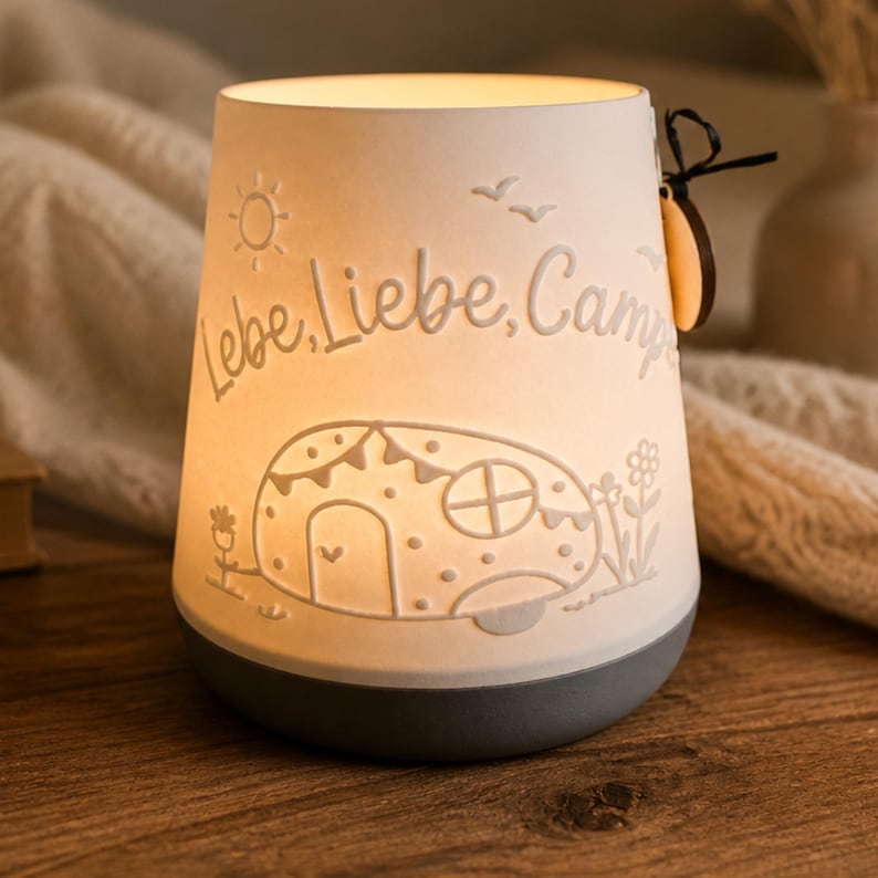 May include: A white ceramic lantern with a grey base, illuminated from within. The surface features embossed designs of a camper, flowers, a sun, and birds, with the text Lebe, Liebe, Campe. A small wooden tag is attached to the side with a black string.