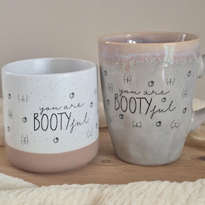 Könnte beinhalten: Zwei Keramikbecher mit dem Schriftzug "you are BOOTYful" in schwarzer Schrift. Ein Becher ist weiß mit rosa Boden und schwarzen Sprenkeln. Der andere ist grau mit Henkel und hellbraunem Rand. Beide Becher haben kleine grafische Illustrationen.