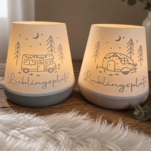 May include: Two illuminated, tapered lamps with a warm glow. Each lamp features a cutout design of a camper, pine trees, and a crescent moon, with the German text "Lieblingsplatz" written in cursive script across the front of the off-white shades.