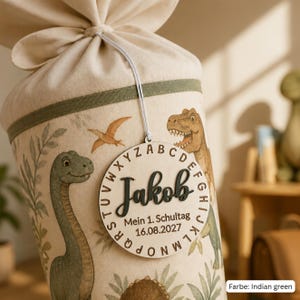 May include: A cream-colored fabric cone with a drawstring top, decorated with dinosaur illustrations and green trim. A round wooden tag hangs from the top, featuring the name Jakob, the alphabet, and the text Mein 1. Schultag 16.08.2027.