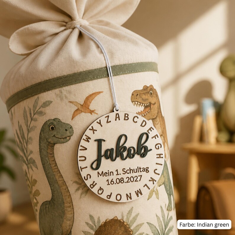 May include: A cream-colored fabric cone with a drawstring top, decorated with dinosaur illustrations and green trim. A round wooden tag hangs from the top, featuring the name Jakob, the alphabet, and the text Mein 1. Schultag 16.08.2027.