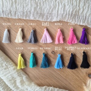May include: Thirteen small fabric tassels arranged in two rows, each featuring a gold-tone metal jump ring at the top. The colors include white, cream, gray, light pink, pink, magenta, purple, yellow, turquoise, petrol, light blue, navy, and black.
