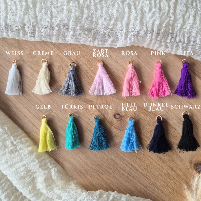 May include: Thirteen small fabric tassels arranged in two rows, each featuring a gold-tone metal jump ring at the top. The colors include white, cream, gray, light pink, pink, magenta, purple, yellow, turquoise, petrol, light blue, navy, and black.
