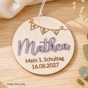 May include: A round wooden ornament featuring a laser-engraved bunting banner with the letters A, B, and C. The name Mathea is displayed in raised, light purple script lettering. Below, the text reads Mein 1. Schultag and 16.08.2027.