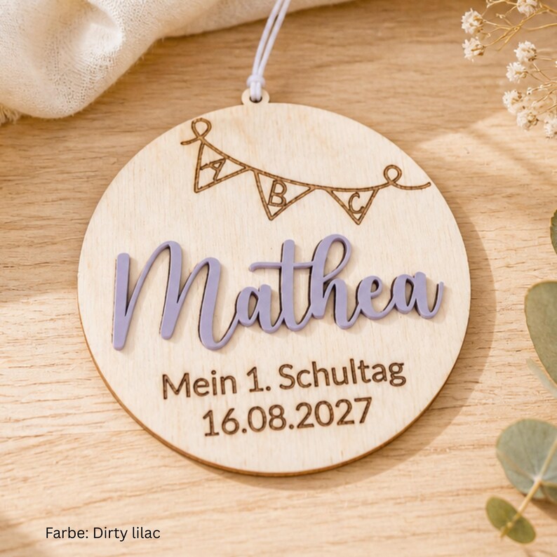 May include: A round wooden ornament featuring a laser-engraved bunting banner with the letters A, B, and C. The name Mathea is displayed in raised, light purple script lettering. Below, the text reads Mein 1. Schultag and 16.08.2027.