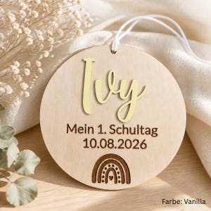 May include: A circular light-colored wooden ornament featuring the name Ivy in gold script lettering. Below the name, the text reads Mein 1. Schultag 10.08.2026, followed by a small engraved rainbow icon. A white string is attached to the top.