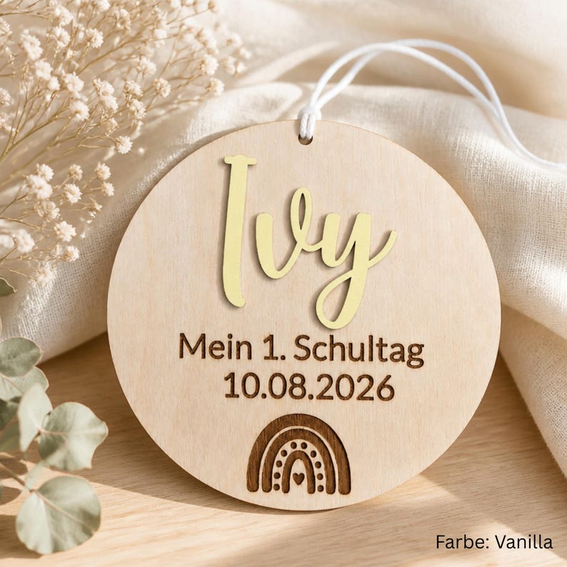 May include: A circular light-colored wooden ornament featuring the name Ivy in gold script lettering. Below the name, the text reads Mein 1. Schultag 10.08.2026, followed by a small engraved rainbow icon. A white string is attached to the top.