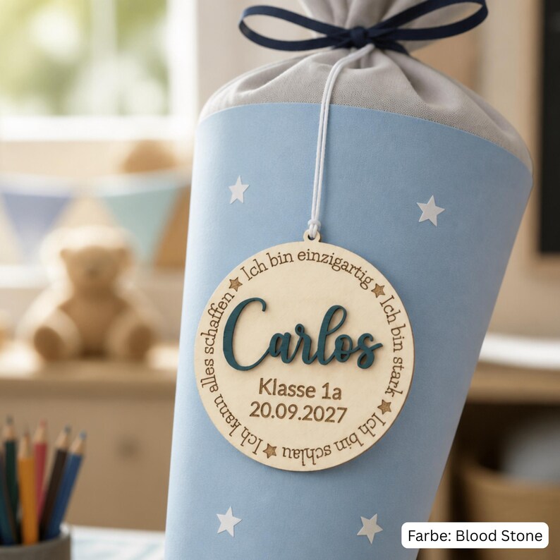 May include: A light blue school cone with white stars and a grey fabric top tied with a dark blue ribbon. A round wooden ornament hangs from the front, featuring the name Carlos in dark blue, plus the text Klasse 1a, 20.09.2027, and German phrases.