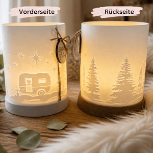 May include: Two cylindrical white lamps with warm internal lighting. The left lamp features a caravan and star cutout design with a small wooden tag labelled "Lieblingsplatz". The right lamp displays a forest of pine trees in silhouette.