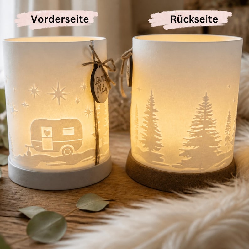 May include: Two cylindrical white lamps with warm internal lighting. The left lamp features a caravan and star cutout design with a small wooden tag labelled "Lieblingsplatz". The right lamp displays a forest of pine trees in silhouette.