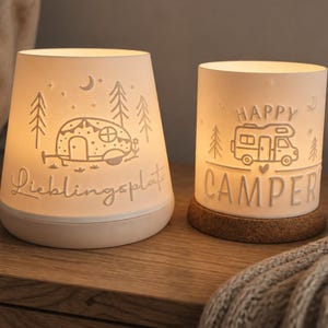 May include: Two illuminated white ceramic lanterns with cut-out designs. The left lantern features a camper trailer and the text "Lieblingsplatz". The right lantern features a motorhome, the text "HAPPY CAMPER", and a cork base.