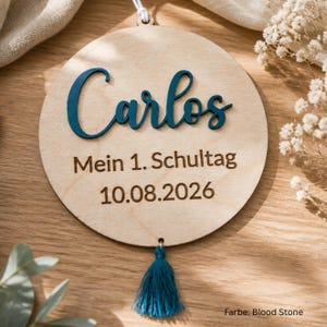 May include: A circular light-colored wooden plaque featuring the name Carlos in raised dark teal script lettering. Below the name, the text Mein 1. Schultag and the date 10.08.2026 are engraved. A dark teal tassel hangs from the bottom of the plaque.