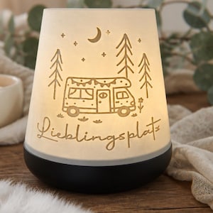 May include: A conical white ceramic lamp with a black base, featuring an illuminated cutout design of a camper van, pine trees, a crescent moon, and stars. The word "Lieblingsplatz" is etched in cursive script across the front of the lamp.