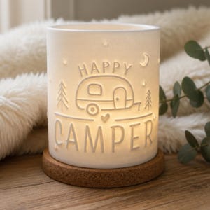 May include: A cylindrical white porcelain tea light holder featuring a cutout design of a camper trailer, pine trees, stars, a crescent moon, and a small heart. The text HAPPY CAMPER is embossed on the front, resting on a circular cork base.