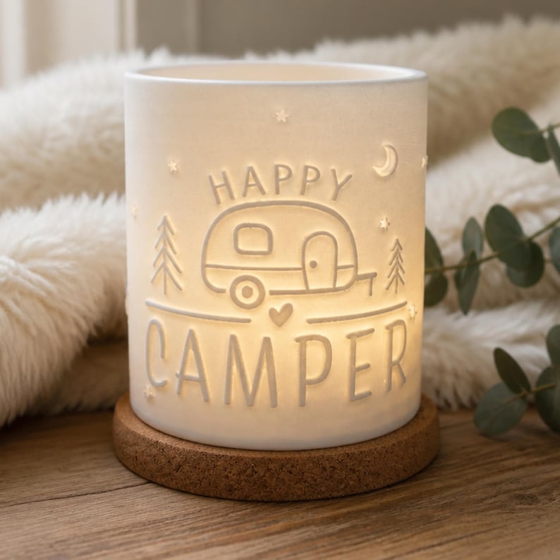 May include: A cylindrical white porcelain tea light holder featuring a cutout design of a camper trailer, pine trees, stars, a crescent moon, and a small heart. The text HAPPY CAMPER is embossed on the front, resting on a circular cork base.