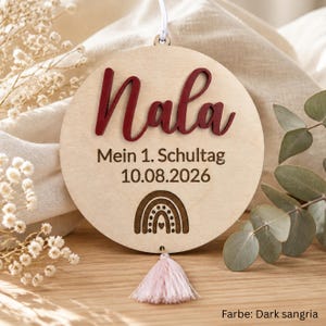 May include: A circular light-colored wooden ornament featuring the name Nala in raised dark red script lettering. Below the name, the text reads Mein 1. Schultag 10.08.2026 with a small engraved rainbow icon. A light pink tassel hangs from the bottom.