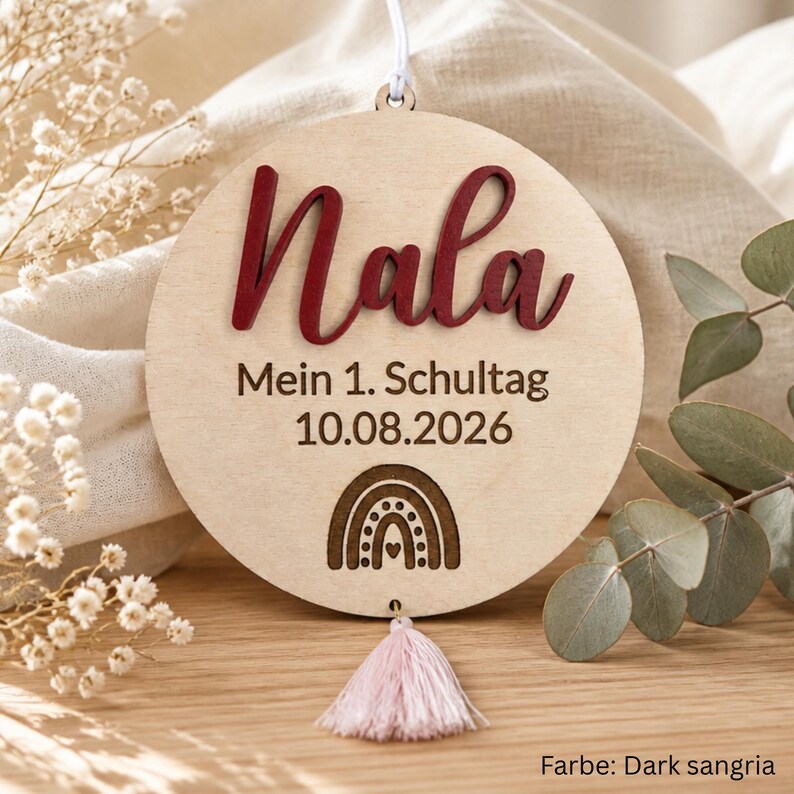 May include: A circular light-colored wooden ornament featuring the name Nala in raised dark red script lettering. Below the name, the text reads Mein 1. Schultag 10.08.2026 with a small engraved rainbow icon. A light pink tassel hangs from the bottom.
