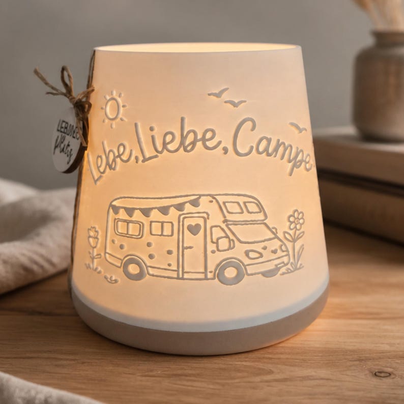 May include: A white ceramic lantern with a glowing interior, featuring an embossed camper van, flowers, birds, and a sun. The text "Lebe, Liebe, Campe" is engraved on the front. A small round wooden tag with the text "LIEBLINGS PLATZ" is attached.