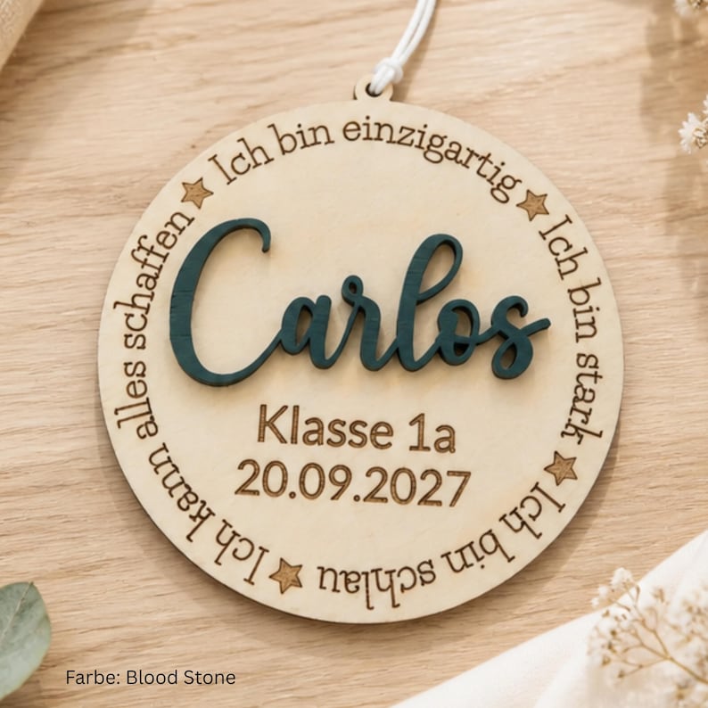 May include: A circular wooden ornament featuring the name Carlos in dark teal 3D lettering. The wood surface is engraved with the text Klasse 1a, 20.09.2027, and the German phrases Ich bin einzigartig, Ich bin stark, Ich bin schlau, Ich kann alles schaffen.