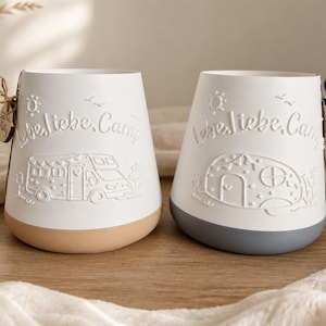 May include: Two white ceramic vessels with embossed designs featuring the text Lebe, liebe, campe. One vessel displays a camper van and has a tan base, while the other shows a rounded travel trailer and has a grey base, both with sun and bird motifs.