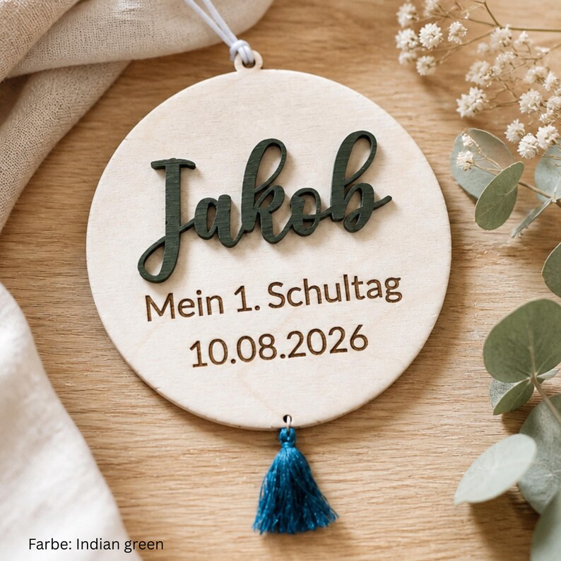 May include: A circular light-colored wooden ornament featuring the name Jakob in dark green raised script lettering. Below the name, the text reads Mein 1. Schultag and the date 10.08.2026. A small blue tassel hangs from the bottom of the wooden disc.