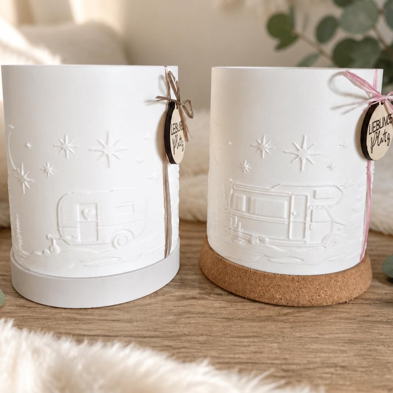 May include: Two white cylindrical candle holders with embossed designs of a travel trailer and a motorhome under a starry sky. Each holder features a small wooden tag tied with string that reads "Lieblings Platz" and sits on a base.