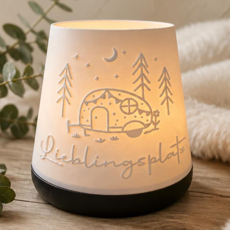 May include: A white porcelain tea light holder with a black base, featuring an illuminated embossed design of a camper trailer, pine trees, stars, and a crescent moon. The word "Lieblingsplatz" is embossed in cursive script on the front surface.