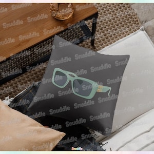 May include: A square, dark gray decorative pillow featuring a stylized illustration of light green sunglasses with dark lenses. The sunglasses have a gold-colored accent on the temple. The pillow is on a white and beige surface.