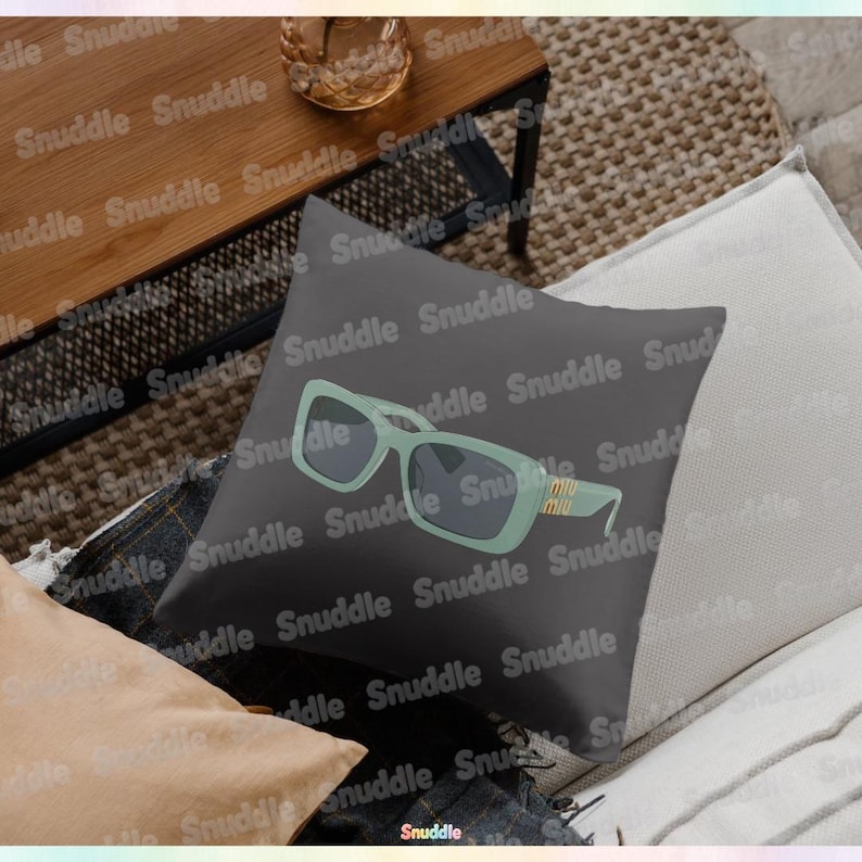 May include: A square, dark gray decorative pillow featuring a stylized illustration of light green sunglasses with dark lenses. The sunglasses have a gold-colored accent on the temple. The pillow is on a white and beige surface.
