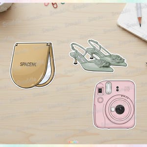 May include: A collection of stickers featuring a tan makeup mirror with the text "SPACENK", a pair of sage green heels with floral accents, and a pink Instax Mini 12 camera.