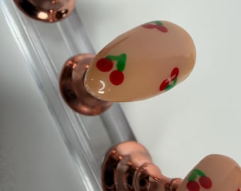 Op de afbeelding: Close-up van een set kunstnagels met een nude basis en kersendesigns. De nagels zijn ovaal van vorm en versierd met kleine rode kersen en groene blaadjes. De nagels zijn bevestigd aan een koperkleurige standaard.