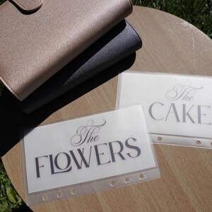 May include: A stack of three notebooks in gold, gray, and black, with a button closure on the top notebook. Two clear plastic sleeves with the words "The FLOWERS" and "The CAKE" printed in elegant script are also visible.
