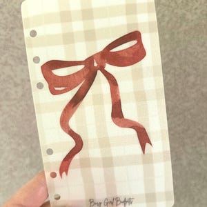 May include: A planner insert with a watercolor-style red bow design on a beige and white gingham background. The insert has three holes for binding and the text "Busy Girl Budgets" in the bottom right corner.