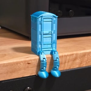 May include: A blue 3D printed model of a portable toilet with legs. The toilet is sitting on a wooden surface.