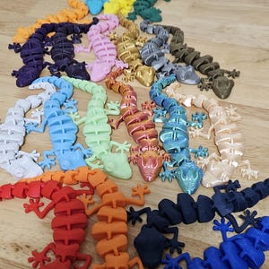 May include: A collection of colorful, articulated lizard figurines in various colors, including blue, yellow, green, and orange. The lizards are arranged on a wooden surface, showcasing their segmented bodies and detailed features.