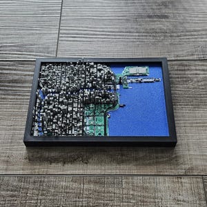 Peut inclure: Une œuvre d'art de paysage urbain en 3D encadrée. L'œuvre présente une ville miniature détaillée avec des bâtiments gris, une zone d'eau bleue et des éléments de circuits imprimés verts. Le cadre est noir, et l'œuvre est posée sur un fond en bois.